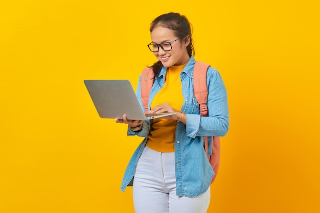 Ritratto di giovane studentessa asiatica sorridente in abiti casual con zaino guardando e-mail in arrivo su laptop isolato su sfondo giallo Istruzione nel concetto di college universitario