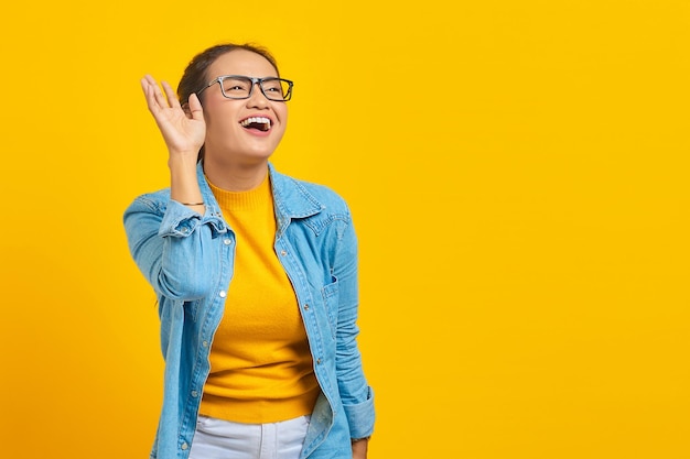 Ritratto di giovane studentessa asiatica sorridente in abiti denim agitando la mano con un amico isolato su sfondo giallo. Persone sincere emozioni concetto di stile di vita