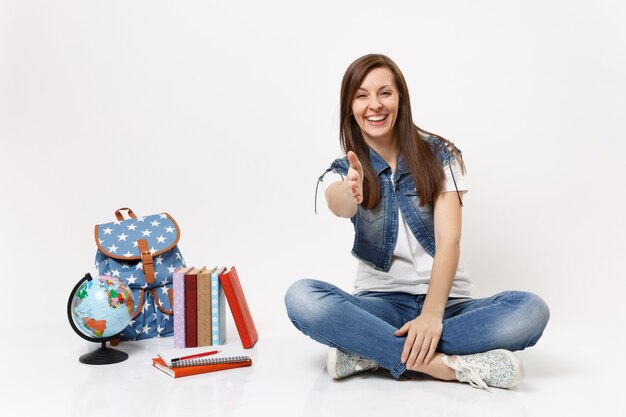 Ritratto di giovane studentessa casuale sorridente che si siede con la mano tesa per il saluto vicino al globo, zaino, libri di scuola isolati