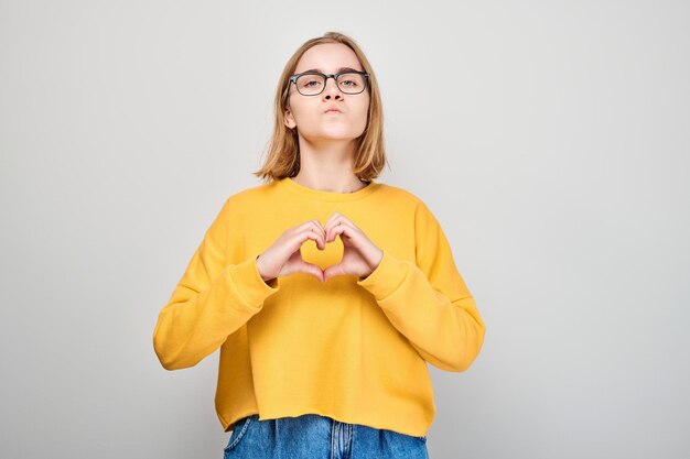 Ritratto di giovane studentessa che invia baci d'aria tenendosi per mano a forma di cuore isolato su sfondo bianco studio