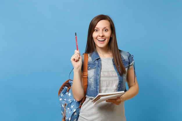 Ritratto di giovane studentessa piuttosto eccitata in abiti in denim con zaino che tiene il taccuino, puntando la matita verso l'alto isolato su sfondo blu. Istruzione nel concetto di college universitario di liceo.