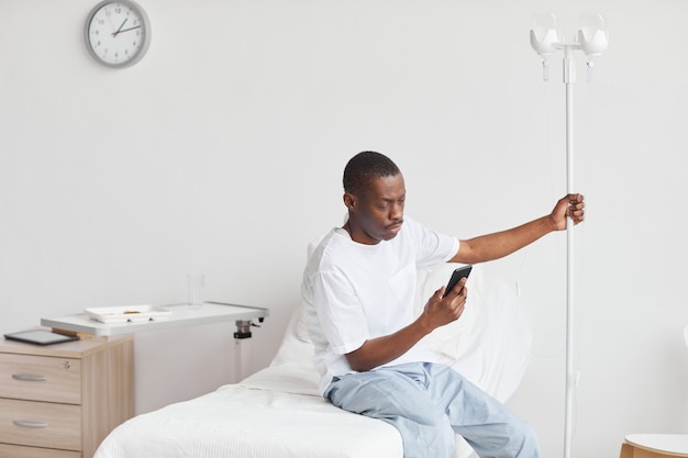 Ritratto di giovane uomo afro-americano che utilizza lo smartphone nella stanza d'ospedale con configurazione di flebo IV, spazio di copia