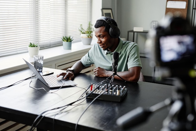 Ritratto di giovane uomo afroamericano che registra podcast in studio e utilizza laptop