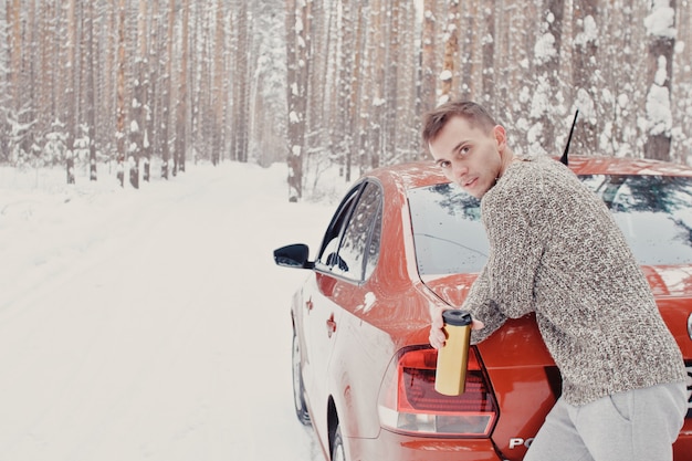 Ritratto di giovane uomo alla moda in abiti grigi con l'auto sulla neve che tiene la valigia e va nella foresta di pini del sentiero. Concetto di viaggio per le vacanze invernali. All'aperto in inverno. Comprensorio sciistico, comprensorio sciistico di montaggio