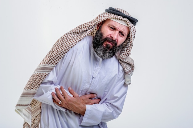 Ritratto di giovane uomo arabo musulmano squezze suo stomaco per avere mal di stomaco, su sfondo bianco.