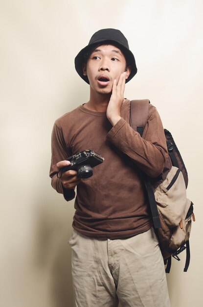 Ritratto di giovane uomo asiatico che indossa un cappello nero che trasporta fotocamera e zaino per il viaggio isolato sullo sfondo