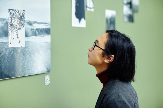 Ritratto di giovane uomo asiatico con i capelli lunghi che indossa occhiali da vista guardando foto in bianco e nero sul muro alla mostra fotografica moderna