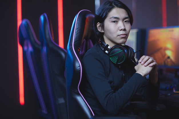 Ritratto di giovane uomo asiatico guardando mentre è seduto nella sedia da gioco in interni scuri di cyber sport, copia dello spazio