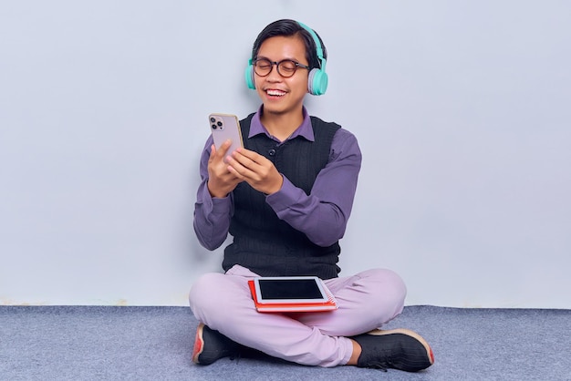 Ritratto di giovane uomo asiatico professionale allegro con gli occhiali che ascolta le cuffie di musica che tengono il telefono cellulare mentre si siede sul pavimento con le gambe incrociate isolate su fondo bianco