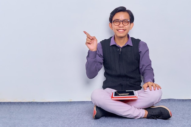 Ritratto di giovane uomo asiatico professionista sorridente con gli occhiali che punta il dito sullo spazio della copia mentre è seduto sul pavimento con le gambe incrociate isolate su sfondo bianco Concetto di tecnologia digitale