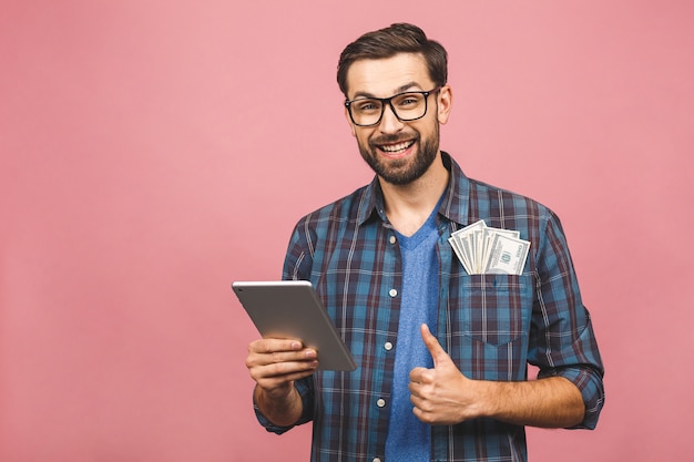 Ritratto di giovane uomo barbuto bello felice hipster nella condizione di camicia a scacchi, tenendo computer tablet e soldi. sfondo rosa. Pollice su.