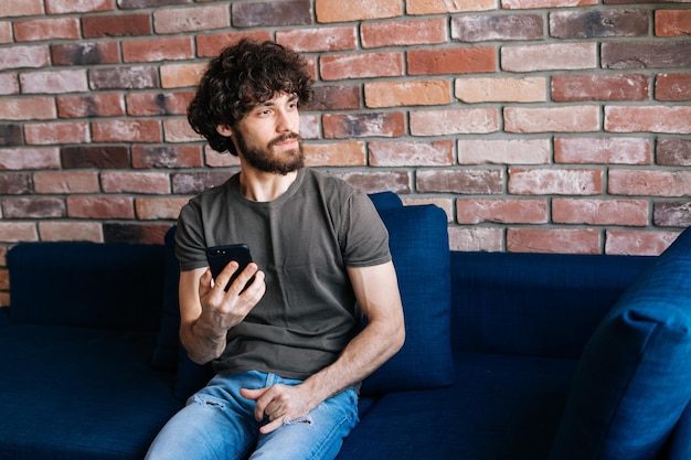 Ritratto di giovane uomo barbuto premuroso che distoglie lo sguardo navigando in internet wireless sul telefono cellulare seduto sul divano di casa