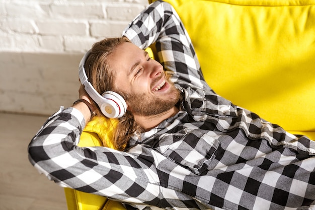 Ritratto di giovane uomo bello che indossa le cuffie ascoltando musica e sdraiato sul divano in appartamento