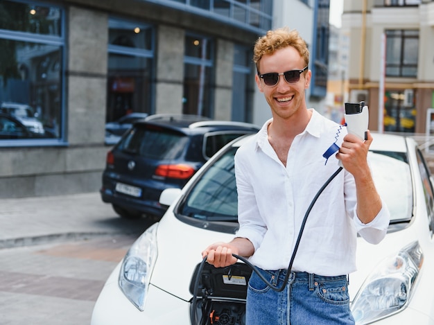 Ritratto di giovane uomo bello in abbigliamento casual, in piedi presso la stazione di ricarica. Concetto di auto elettrica ecologica