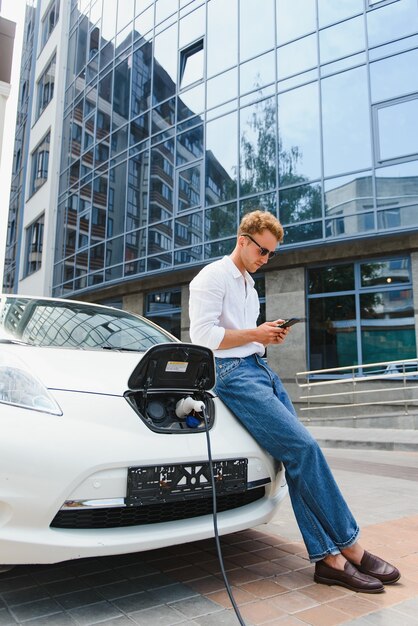 Ritratto di giovane uomo bello in abbigliamento casual, in piedi presso la stazione di ricarica. Concetto di auto elettrica ecologica