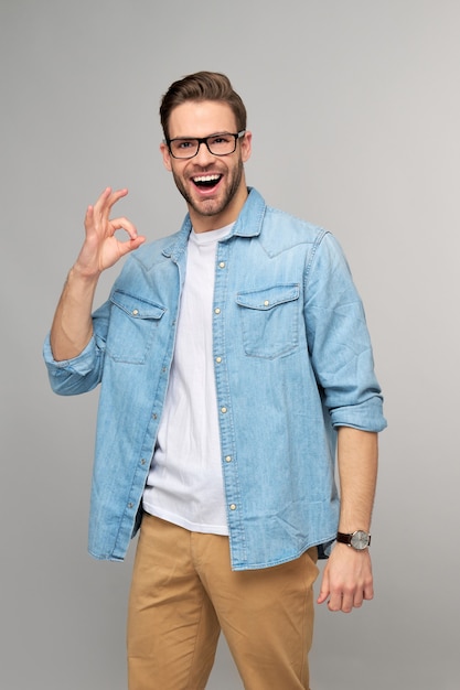 Ritratto di giovane uomo bello in camicia di jeans che mostra il gesto del segno giusto in piedi sopra la parete leggera