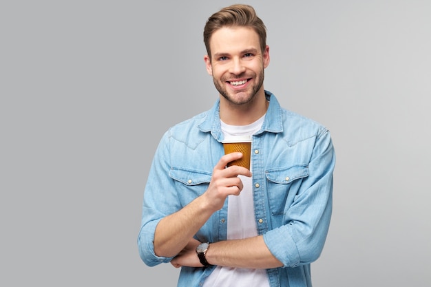 Ritratto di giovane uomo bello in camicia di jeans sopra la parete leggera che tiene tazza di caffè per andare