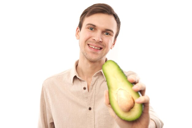 Ritratto di giovane uomo castana caucasico bello che sorride e che mostra mezzo avocado isolato su sfondo bianco studio