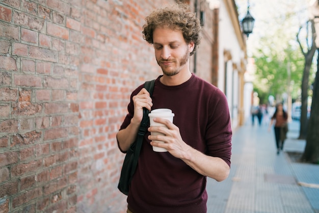 Ritratto di giovane uomo che cammina e che tiene una tazza di caffè all'aperto sulla strada. Concetto urbano.