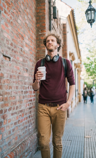 Ritratto di giovane uomo che cammina e che tiene una tazza di caffè all'aperto sulla strada. Concetto urbano.