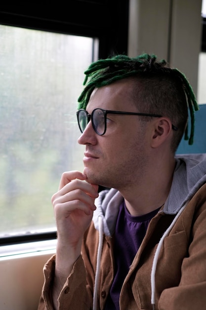 Ritratto di giovane uomo con dreadlocks verdi in occhiali che cavalcano sul treno guardando la natura Primo piano
