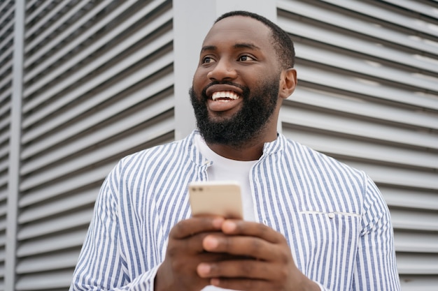 Ritratto di giovane uomo d'affari africano fiducioso che tiene il cellulare, comunicazione, lavoro in linea, in piedi sulla strada