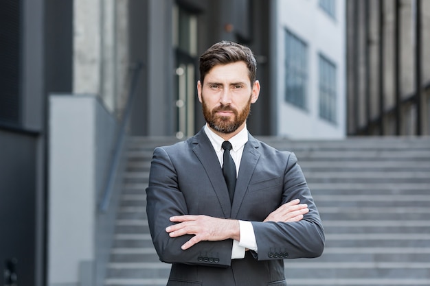 Ritratto di giovane uomo d'affari barbuto caucasico di successo fiducioso che guarda l'obbiettivo Uomo d'affari serio in piedi sullo sfondo urbano dell'edificio per uffici moderno all'aperto Maschio un vestito formale con le braccia incrociate