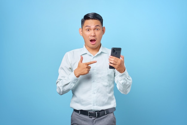 Ritratto di giovane uomo d'affari bello scioccato che punta il dito sullo smartphone su sfondo blu