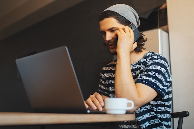 Ritratto di giovane uomo d'affari che ha una conversazione con l'altra persona dallo smartphone; usando il laptop e bevendo caffè.