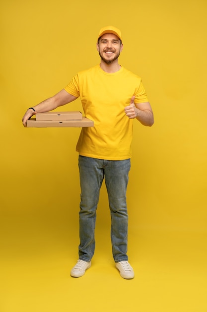 Ritratto di giovane uomo di consegna in uniforme gialla con scatole per pizza isolate sopra la parete gialla