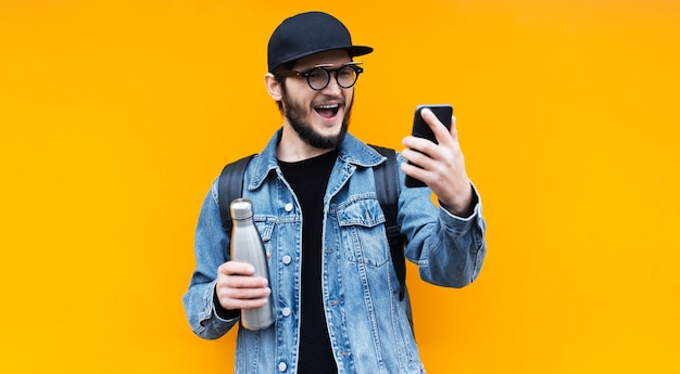 Ritratto di giovane uomo felice hipster sulla parete arancione. Scatta foto selfie su smartphone con la bottiglia termica in acciaio per l'acqua in un'altra mano.