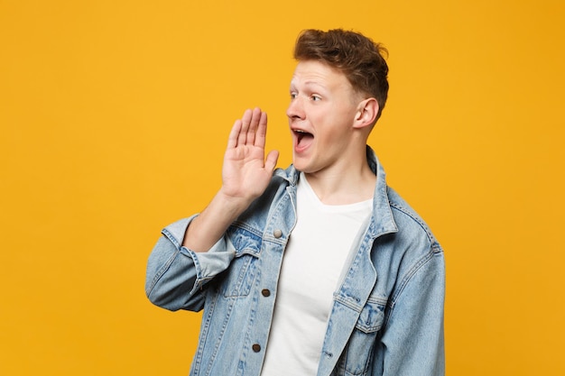 Ritratto di giovane uomo in abiti casual denim che guarda da parte, urlando con il gesto della mano isolato su sfondo giallo arancione in studio. Persone sincere emozioni concetto di stile di vita. Mock up spazio di copia.