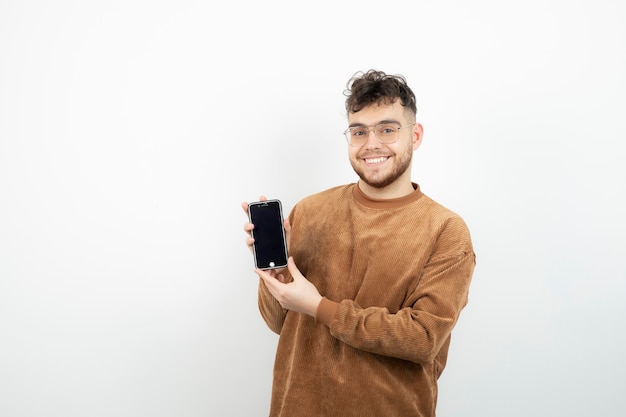 Ritratto di giovane uomo in bicchieri tenendo il cellulare sul muro bianco.