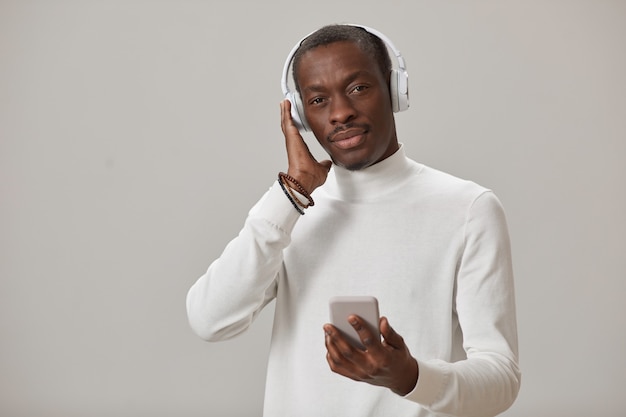 Ritratto di giovane uomo in cuffie senza fili utilizzando il telefono cellulare per ascoltare musica isolata sul muro grigio