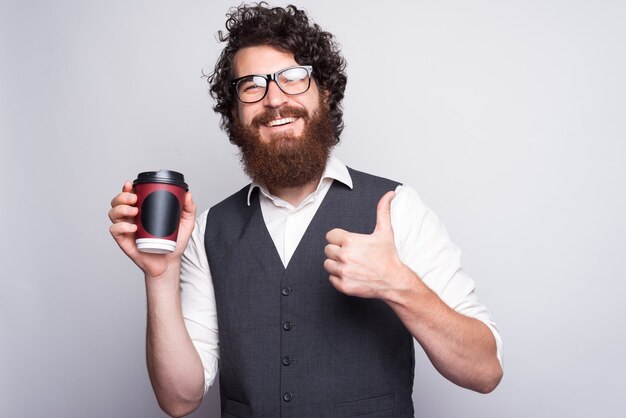 Ritratto di giovane uomo in tuta con gli occhiali e tenendo la tazza di caffè a gom, pollice in alto.