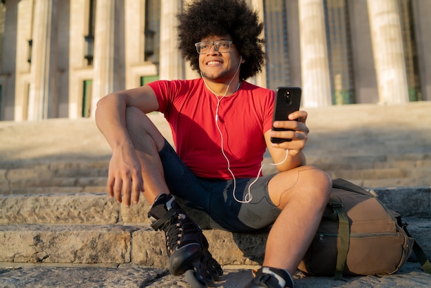 Ritratto di giovane uomo latino utilizzando il suo telefono cellulare e indossando rulli da skate mentre è seduto all'aperto. Concetto di sport