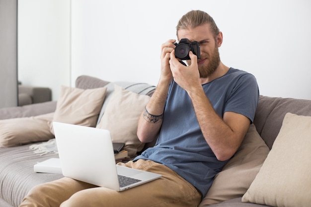 Ritratto di giovane uomo seduto sul divano grigio con laptop sulle ginocchia e fotocamera in mano a casa