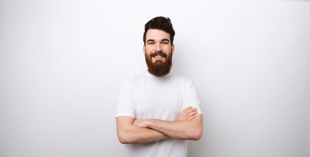 Ritratto di giovane uomo sorridente e bello con la barba con le braccia incrociate in piedi sul muro bianco