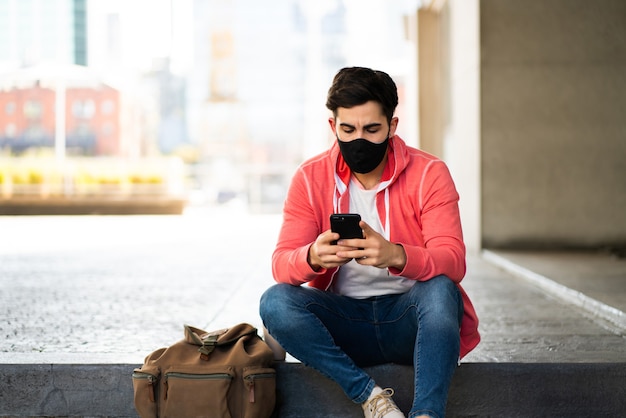 Ritratto di giovane uomo utilizzando il suo telefono cellulare mentre è seduto all'aperto in strada. Uomo che indossa la maschera per il viso. Concetto urbano.