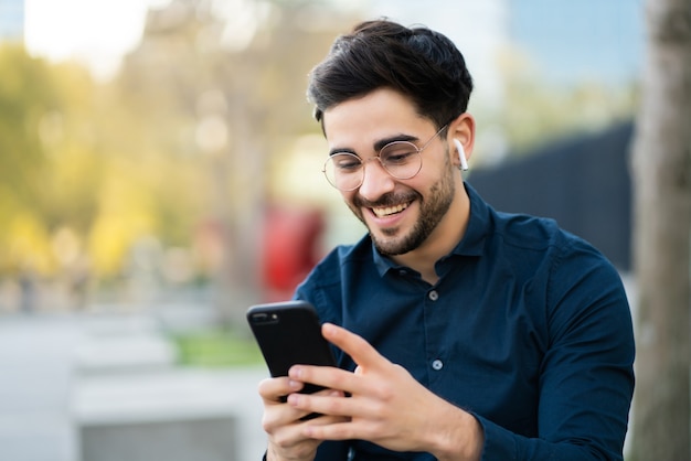 Ritratto di giovane uomo utilizzando il suo telefono cellulare mentre è seduto su una panchina all'aperto. Concetto urbano.