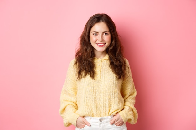 Ritratto di goodlooking giovane donna in maglione tenendo le mani in tasca sorridendo e fissando determinato a porte chiuse in piedi contro il muro rosa