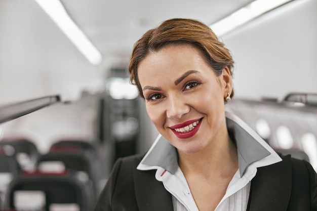 Ritratto di hostess nella cabina passeggeri del jet aereo. Sfocatura degli interni dell'aereo moderno. La donna caucasica sorridente indossa l'uniforme e guarda la telecamera. Aviazione civile commerciale. Concetto di viaggio aereo