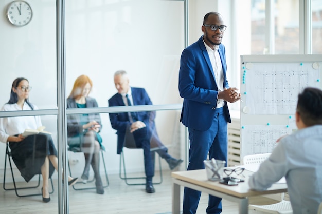 Ritratto di imprenditore afro-americano di successo in piedi dalla lavagna mentre presenta il progetto di design durante la riunione in ufficio, copia ritmo