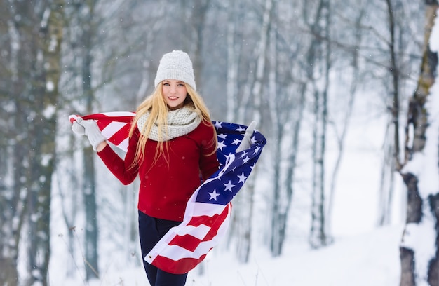 Ritratto di inverno di giovane ragazza teenager con la bandiera di USA Ragazza di modello allegra di bellezza che ride e che si diverte nel parco di inverno