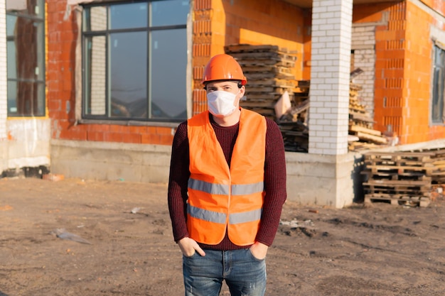 Ritratto di lavoratore edile maschile in maschera medica e tuta sullo sfondo della casa in costruzione