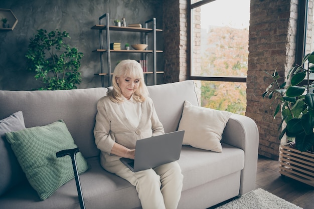 Ritratto di lei bella signora dai capelli grigi amichevole pacifica attraente che si siede sul divano scrivendo e-mail in chat ricerca sito web al loft industriale in stile moderno interni casa al chiuso