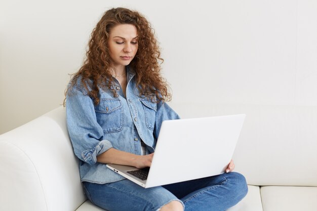 Ritratto di libero professionista concentrato che lavora online a casa, utilizzando la tecnologia moderna, affascinante femmina con lunghi capelli ricci che indossa giacca di jeans, seduto in una stanza accogliente sul comodo divano bianco bianco.