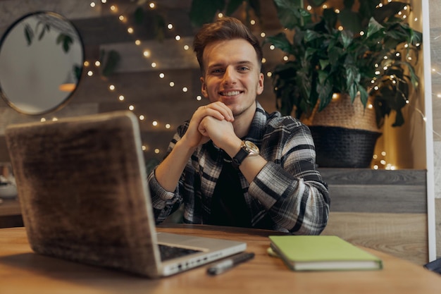 Ritratto di libero professionista maschio sorridente seduto al tavolo nella caffetteria con il laptop e guardando la fotocamera