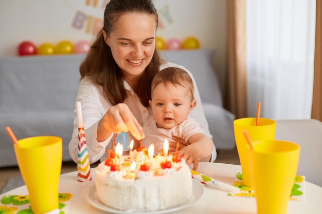 Ritratto di madre e bambino con torta di compleanno seduti a tavola e guardando un delizioso dessert, festeggiando il compleanno del primo bambino, posando a casa con divano e palloncini sullo sfondo.