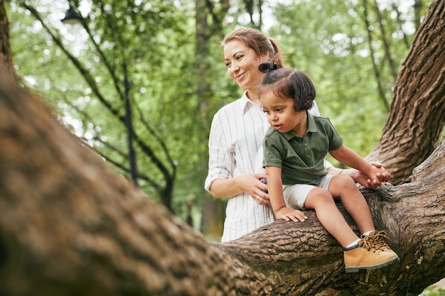 Ritratto di madre felice che gioca con un ragazzo carino nel parco seduto sui rami degli alberi, copia spazio